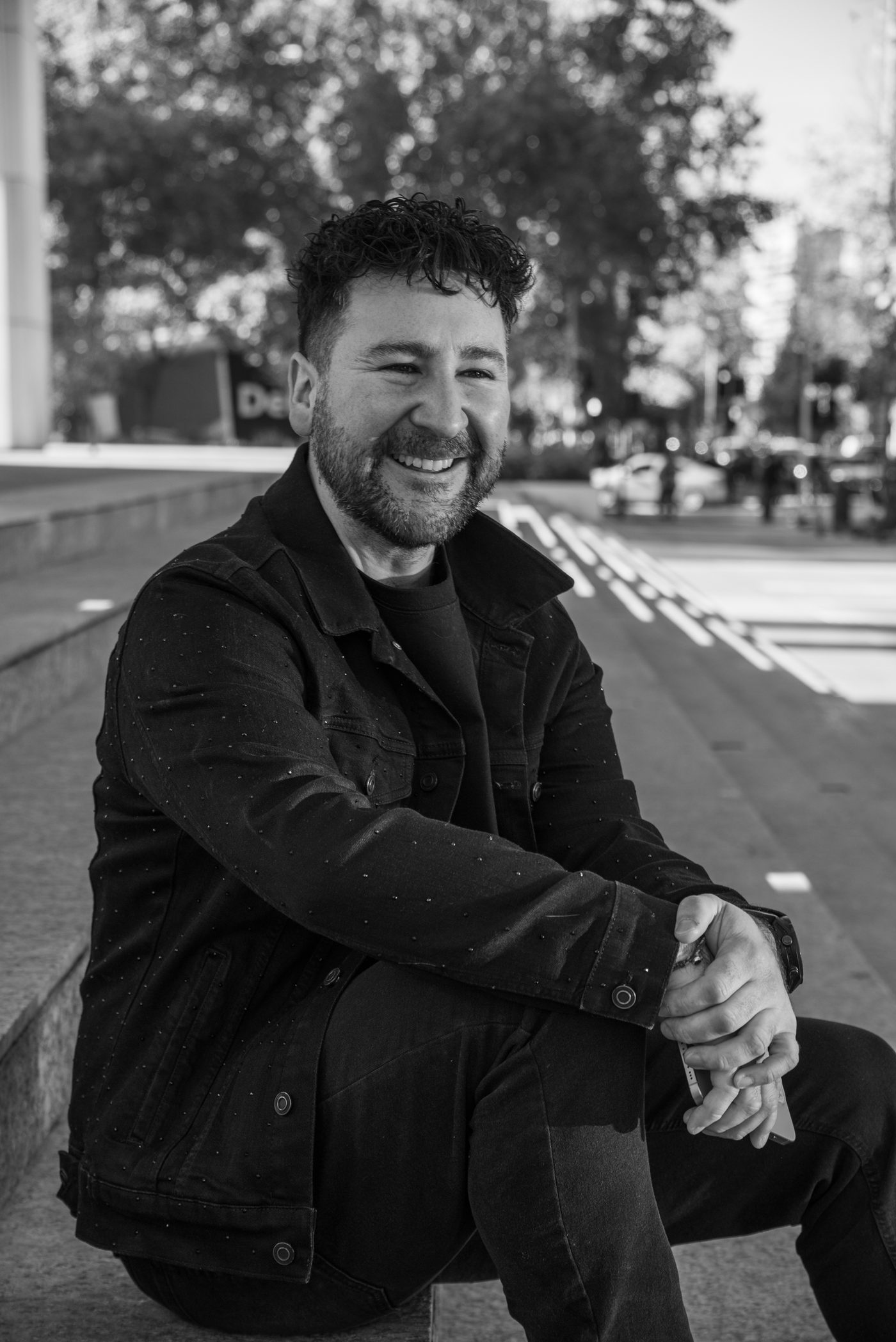 Francisco Sáez sonriendo, sentado en una calle de la ciudad, con chaqueta oscura; foto en blanco y negro.